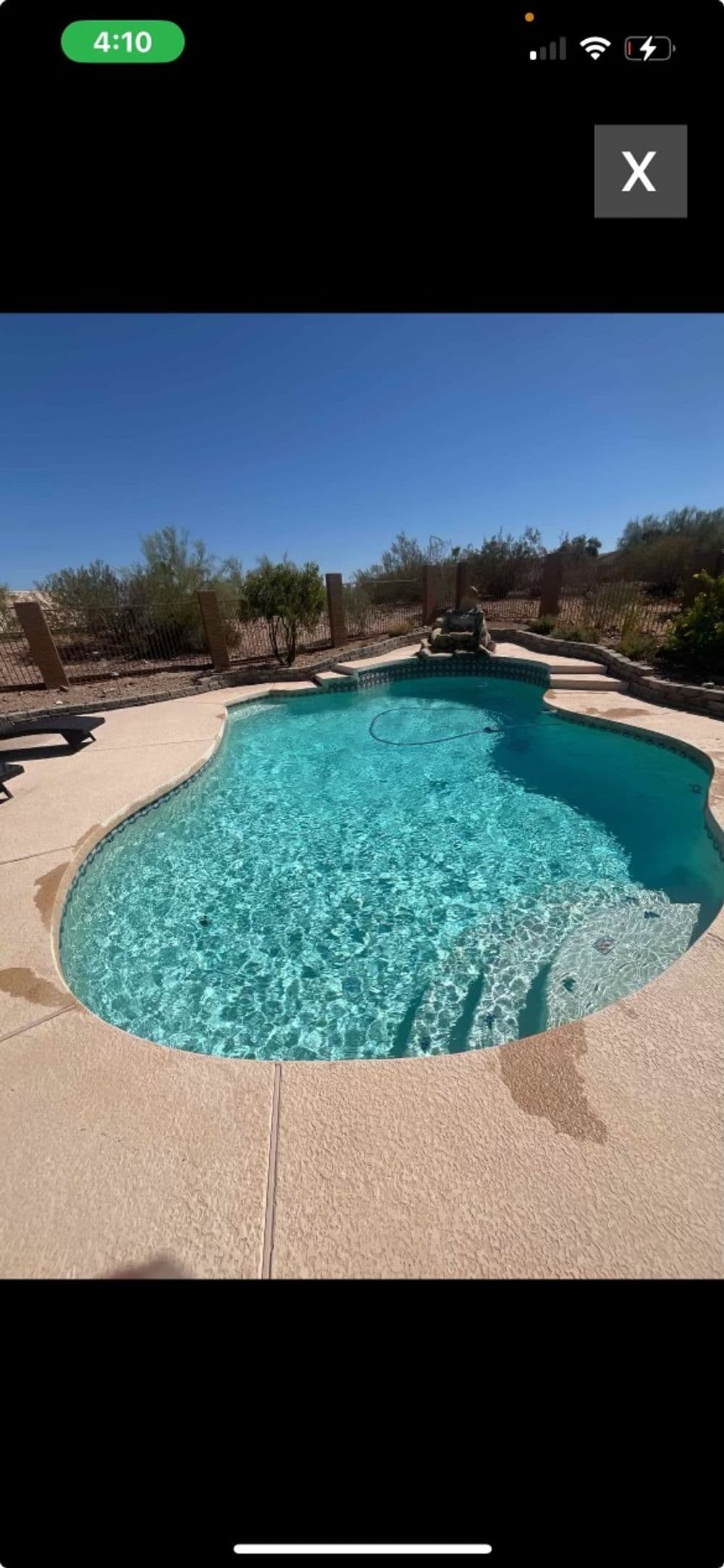 Clear blue swimming pool surrounded by desert landscape and fencing under a sunny sky.