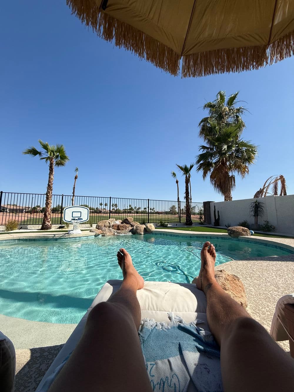 Person relaxing by a pool under a parasol, surrounded by palm trees on a sunny day.